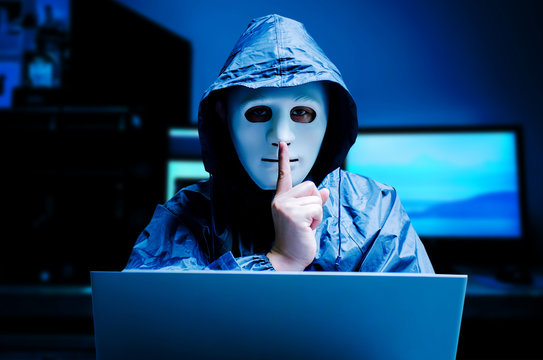 Anonymous computer hacker in white mask and hoodie. Obscured dark face making silence gesture on dark background, Data thief, internet attack, darknet and cyber security concept.
