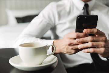Cropped image of successful man holding smartphone and drinking coffee in hotel apartment during business trip