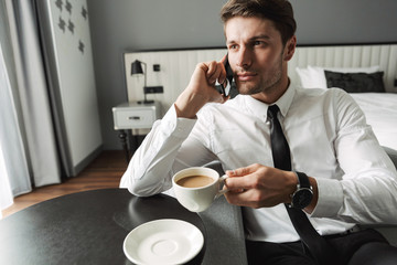Image of masculine young man talking on smartphone and drinking coffee in hotel apartment during...