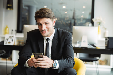 Portrait of successful handsome businessman sitting on armchair with smartphone in hotel hall