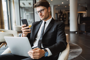 Portrait of beautiful businessman using laptop computer and holding smartphone in hotel hall