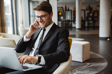 Photo of happy young businessman using laptop computer and talking on smartphone in hotel hall - Powered by Adobe