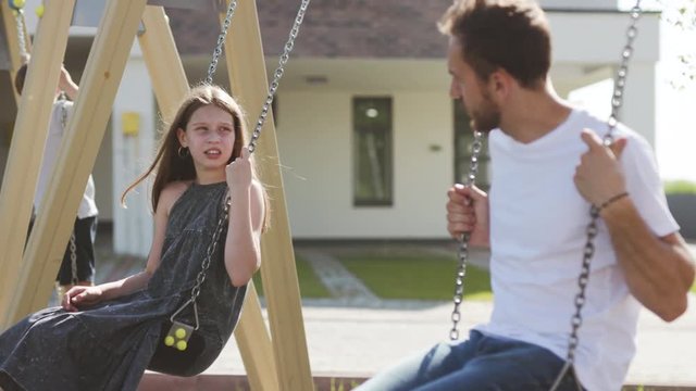 Young family swinging in the playground on sunny day. Upset daughter and father talking together solving problems outdoors in the courtyard.