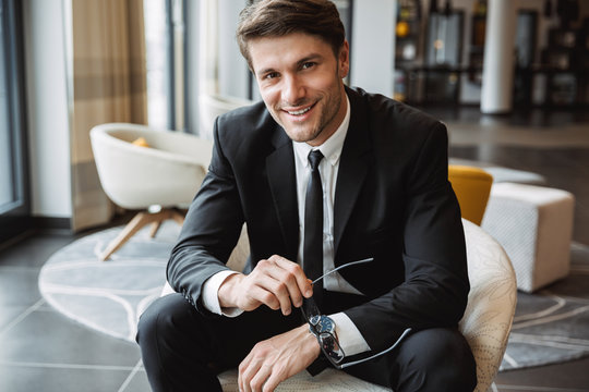 Photo of smiling businessman holding eyeglasses and sitiing on armchair in hotel lobby