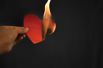 The hands of an Asian woman are holding a red paper heart that is burning on a dark background.