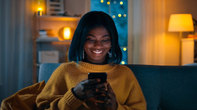 Portrait of Beautiful Black Girl Uses Smartphone while Sitting on a Couch at Home, Her Face is Illuminated with a Screen Light. In the Cute Woman Smiles and Browses Through Internet.