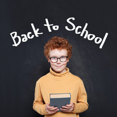 Smiling kid boy holding book on chalkboard background with Back to school text