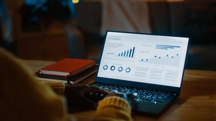 Over the Shoulder Shot: Woman Sitting at Her Desk Works on a Laptop that Shows Statistics and Infographics. Late at Night in Her Living Room Woman Uses Computer.