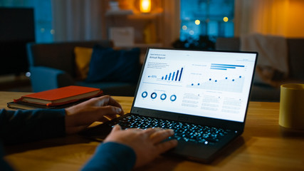 Over the Shoulder Shot: Young Man Sitting at His Desk Works on a Laptop that Shows Statistics and Infographics. Late at Night in His Living Room Man Uses Computer.