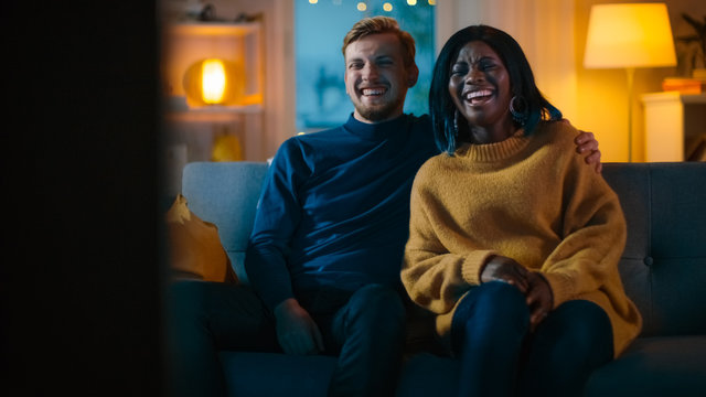 Happy Diverse Young Couple Watching Comedy On TV While Sitting On A Couch, They Laugh And Enjoy Show. Handsome Caucasian Boy And Black Girl In Love Spending Time Together In The Cozy Apartment.