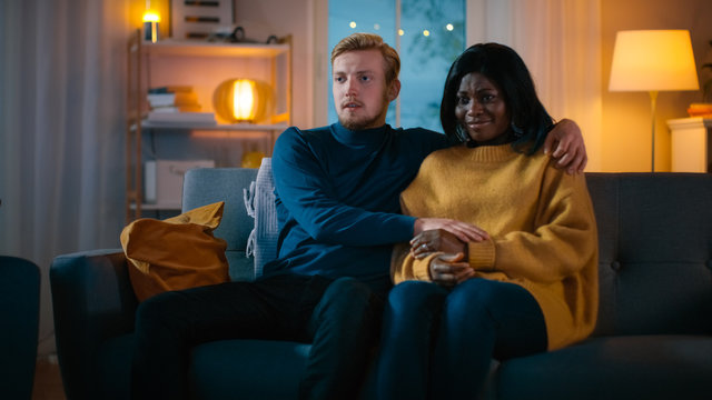 Happy Young Couple Watching Horror Movie on TV while Sitting on a Couch, they got Scared, Girl Covers eyes. Handsome Caucasian Boy and Black Girl in Love Spending Time Together.