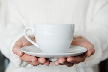 Fototapeta na wymiar Woman in white knitted sweater holding cup of coffee. Female hands with cup of tea. Close up