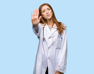 Redhead doctor woman making stop gesture denying a situation that thinks wrong on isolated blue...