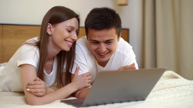 Young happy caucasian young man and woman lying on the bed, talking and using laptop in bright bedroom