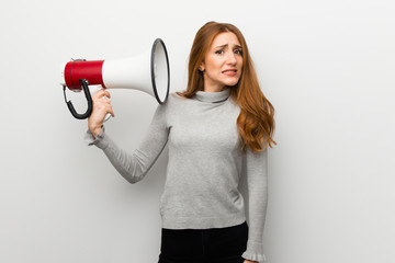 Redhead girl over white wall taking a megaphone that makes a lot of noise