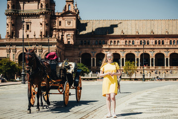 Fototapeta premium Summer outdoor portrait of young beautiful woman in yellow dress. Cheerful blond girl walking between old stone houses in city square. Enjoying vacation in Portugal. Travel concept