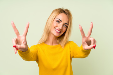 Young blonde woman over isolated green background smiling and showing victory sign