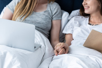 partial view of two lesbians with laptop and book holding hands while lying under blanket in bed
