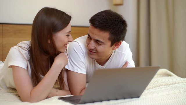 Young couple of girls and guy lying on bed in front of laptop in light bedroom. Pretty young woman flirts with her boyfriend and tickles his face gently with her hair