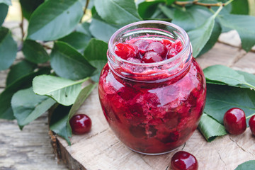 Bank with homemade cherry jam on a wooden background near the berries and leaves.