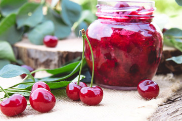 Bank with homemade cherry jam on a wooden background near the berries and leaves.