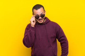 Colombian man with sweatshirt over yellow wall with glasses and smiling