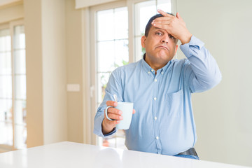 Middle age man drinking coffee in the morning at home stressed with hand on head, shocked with shame and surprise face, angry and frustrated. Fear and upset for mistake.