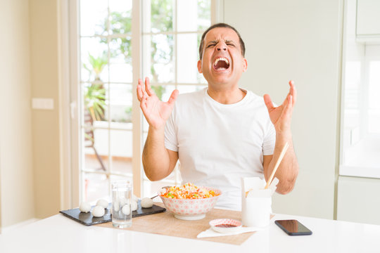 Middle age man eating asian food with chopsticks at home celebrating mad and crazy for success with arms raised and closed eyes screaming excited. Winner concept
