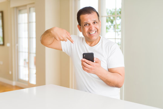 Middle age man using smartphone at home with surprise face pointing finger to himself
