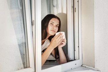 young girl with cup of coffee in her hands looks out open window of house