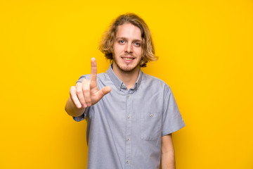Blonde man over isolated yellow background showing and lifting a finger
