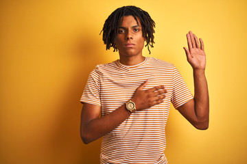Afro man with dreadlocks wearing striped t-shirt standing over isolated yellow background Swearing...