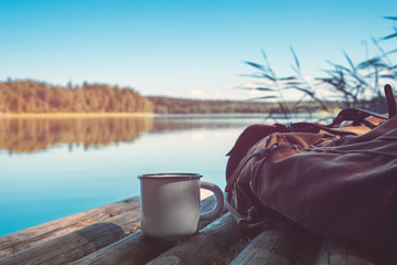 Enameled cup of coffee or tea, backpack of traveller on wooden pier on summer tranquil lake.