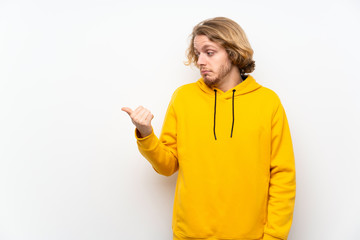 Blonde man with  sweatshirt over white wall unhappy and pointing to the side