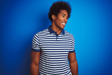 African american man with afro hair wearing striped polo standing over isolated blue background looking away to side with smile on face, natural expression. Laughing confident.
