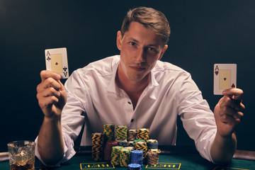 Handsome emotional man is playing poker sitting at the table in casino against a white spotlight.