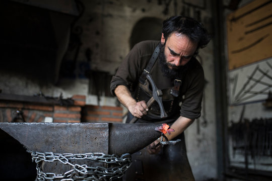 Man in leather apron hitting piece of hot metal with heavy hammer
