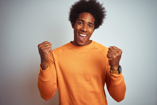 Young american man with afro hair wearing orange sweater over isolated white background celebrating surprised and amazed for success with arms raised and open eyes. Winner concept.