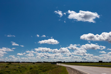 Cumulus clouds in the blue sky over a field with a highway. Sky pattern.