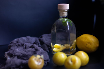 Plum tincture in a square bottle. Closed wooden stopper. Near yellow plums and a big lemon. All on a black background.