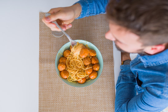 Handsome man eating pasta with meatballs and tomato sauce at home while smiling at the camera