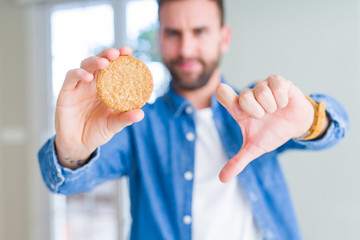 Handsome man eating healthy whole grain biscuit with angry face, negative sign showing dislike with thumbs down, rejection concept