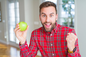 Handsome man eating fresh healthy green apple screaming proud and celebrating victory and success very excited, cheering emotion