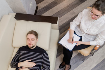 Young man at the psychotherapist, lying on couch, emotionally speaking, discussing with psychotherapist his problems, practitioner making some notes