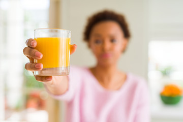 Young african american woman driking orange juice at home with a confident expression on smart face thinking serious