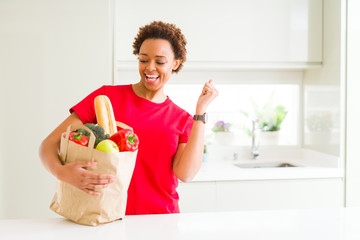 Young african american woman holding paper bag full of fresh groceries screaming proud and...