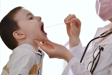 The child sits with his mouth wide open, and the doctor holds a throat examination with a spatula.