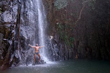 Happy young man relaxing under tropical waterfall with arms up raised in freedom. Health and relaxation.