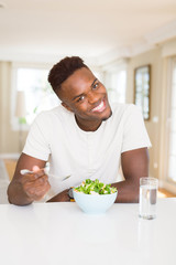 Fototapeta na wymiar Handsome african young man eating a healthy vegetable salad using a fork to eat lettuce, happy and smiling sitting on the table