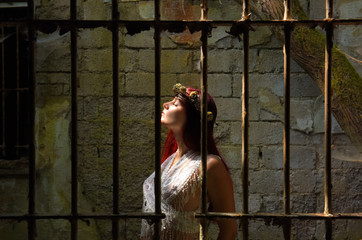 A young woman is trapped inside a jail ruin. She is  dressed in white and wears a thorn wreath on...